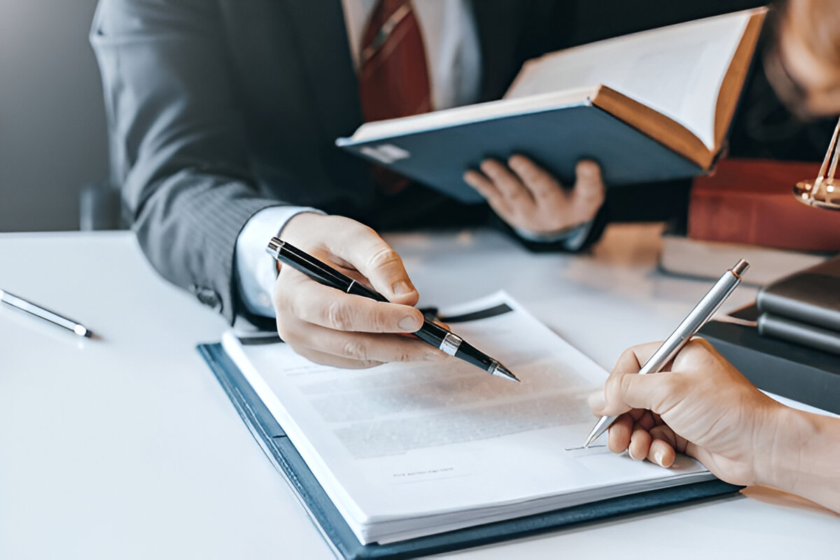 Business professional in suit signing documents with pen while another person holds open book, with papers and desk accessories visible on white table